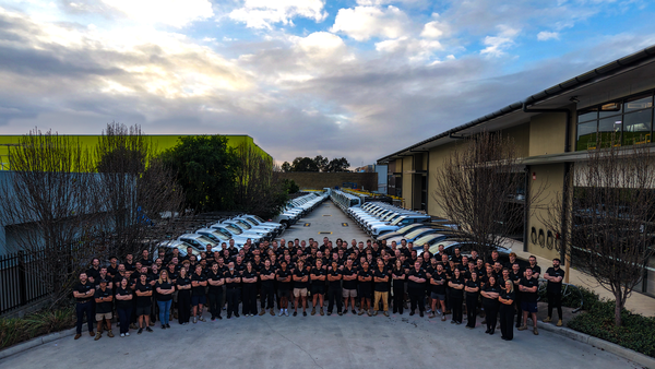 A large group of people stand in rows in front of multiple parked cars on a concrete lot between two buildings under a partly cloudy sky.