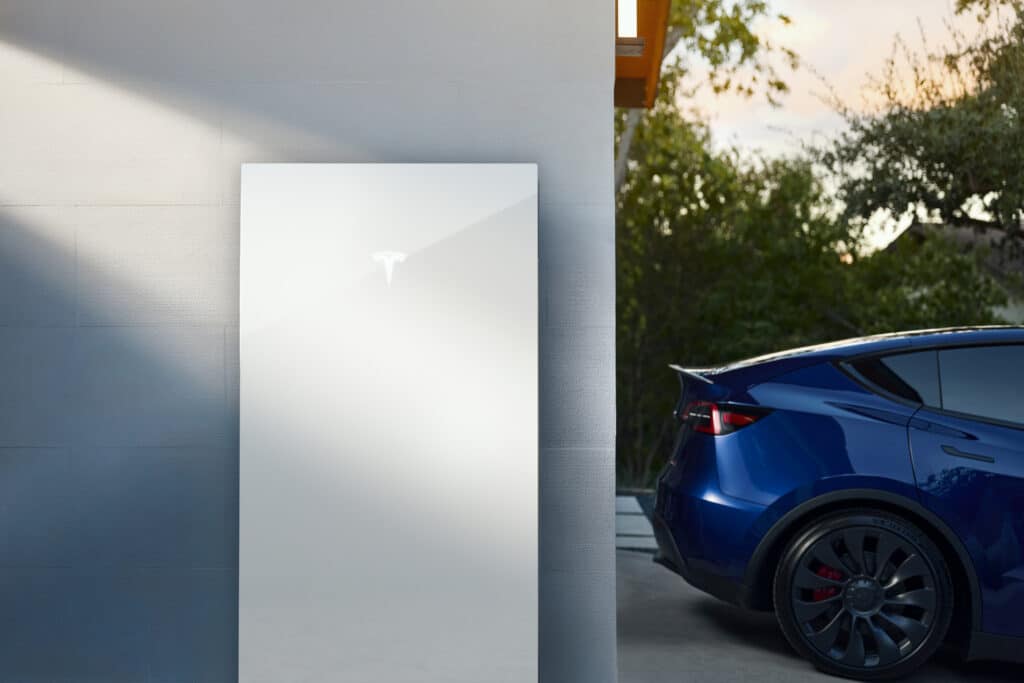 A Tesla Powerwall is mounted on an exterior wall near a parked blue Tesla vehicle in a modern outdoor setting.