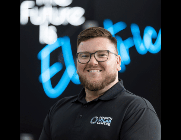 A smiling man in a black polo shirt standing in front of a blurred sign that reads "the future is electric.