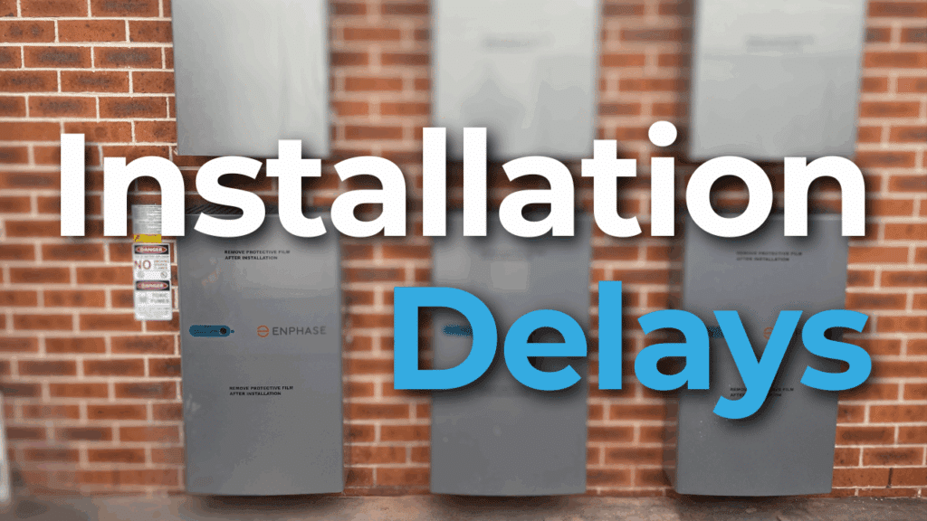 Three Enphase battery storage units mounted on a brick wall with a sign indicating "Installation Delays" overlaying the image.